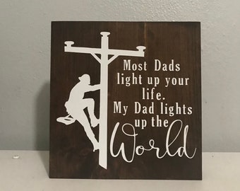 May include: A brown wooden sign with white text that reads "Most Dads light up your life. My Dad lights up the world". A silhouette of a person climbing a utility pole is on the left side of the sign.