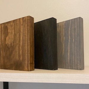 May include: Three wooden blocks in different shades of brown, black, and gray. The blocks are arranged side by side on a white shelf.