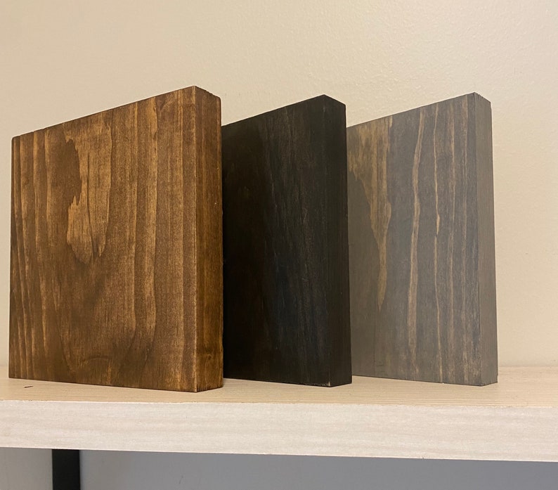 May include: Three wooden blocks in different shades of brown, black, and gray. The blocks are arranged side by side on a white shelf.