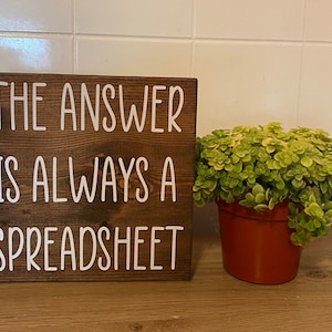 May include: A brown wooden sign with white text that reads "The answer is always a spreadsheet".