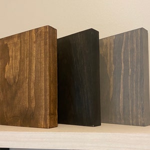 May include: Three wooden blocks in different shades of brown, black, and gray. The blocks are all the same size and shape, and they are arranged in a row from left to right.