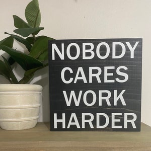 May include: Black wooden sign with white text that reads "Nobody Cares Work Harder".