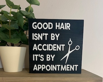 Puede incluir: Un letrero de madera negro con texto blanco que dice "Good hair isn't by accident it's by appointment". Un par de tijeras blancas se representa en el letrero.