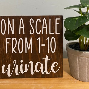May include: A brown wooden sign with white text that reads "On a scale from 1-10 urinate".