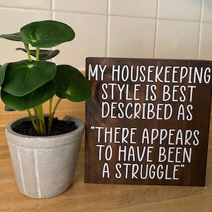 May include: A brown wooden sign with white text that reads "My housekeeping style is best described as "There appears to have been a struggle."