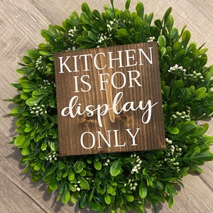 May include: A square, dark brown wooden sign with white lettering that reads "KITCHEN IS FOR display ONLY." The sign is centered on a green wreath with small white flowers.