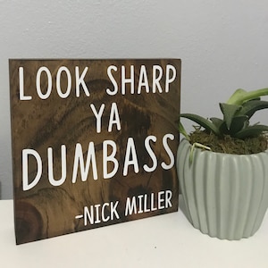 May include: A wooden sign with the text "Look Sharp Ya Dumbass - Nick Miller" in white lettering on a brown background.