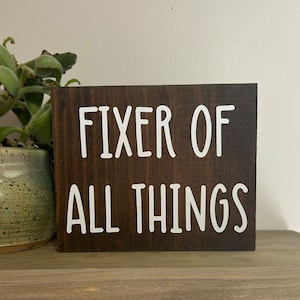 May include: A dark brown wooden sign with white text that reads "Fixer of All Things".