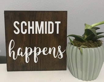 May include: A wooden sign with the text "Schmidt happens" in white lettering on a dark brown background. The sign is sitting on a table next to a small green potted plant.