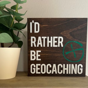 May include: A brown wooden sign with white text that reads "I'd rather be geocaching". A green compass symbol is on the right side of the sign.