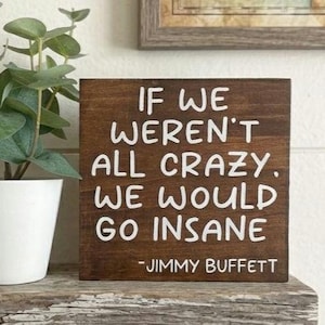 May include: A square, wooden sign with the quote "IF WE WEREN'T ALL CRAZY, WE WOULD GO INSANE" in white lettering. The sign is a dark brown color and includes the name "JIMMY BUFFETT" at the bottom. A potted plant is to the left.