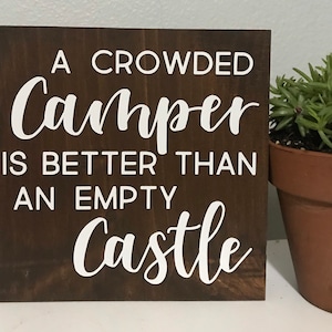 May include: A wooden sign with the text "A crowded camper is better than an empty castle" in white lettering on a dark brown background.