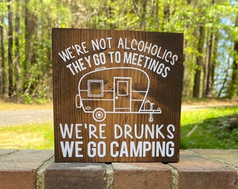 May include: A brown wooden sign with white text that reads "We're not alcoholics they go to meetings. We're drunks we go camping." A white silhouette of a camper trailer is in the middle of the sign.