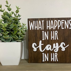 May include: A brown wooden sign with white text that reads "What happens in HR stays in HR".
