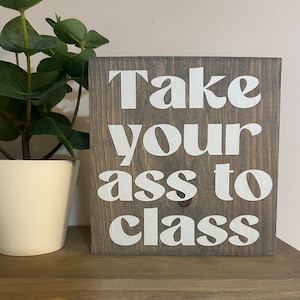 May include: A square, wooden sign with the phrase "Take your ass to class" in white, bold letters. The sign has a gray-brown wood grain finish. A potted plant with green leaves sits to the left of the sign.