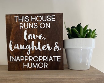 May include: A brown wooden sign with white text that reads "This house runs on love, laughter & inappropriate humor".