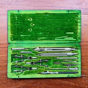 May include: An open, vintage drafting set in a green velvet-lined case. The set includes various silver-colored compasses, dividers, and other drawing tools. The case is rectangular and sits on a wooden surface.