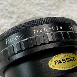 K&ouml;nnte beinhalten: Nahaufnahme einer schwarzen Bicar-Fischaugenlinse. Auf der Linse stehen die W&ouml;rter "fish-eye lens" und "Bicar" in wei&szlig;er Schrift. Ein goldener Aufkleber mit dem Wort "PASSED" ist ebenfalls sichtbar.