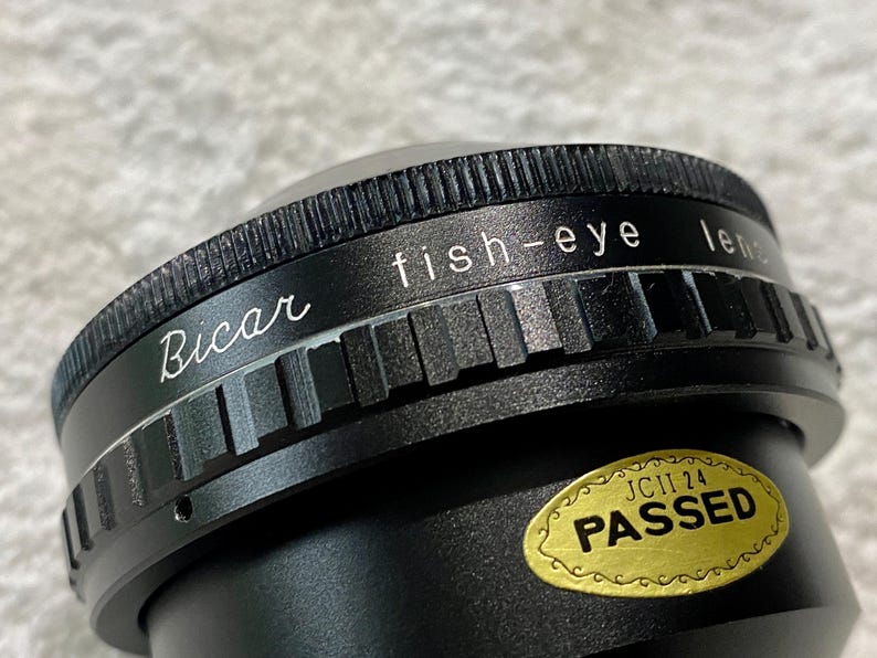 K&ouml;nnte beinhalten: Nahaufnahme einer schwarzen Bicar-Fischaugenlinse. Auf der Linse stehen die W&ouml;rter "fish-eye lens" und "Bicar" in wei&szlig;er Schrift. Ein goldener Aufkleber mit dem Wort "PASSED" ist ebenfalls sichtbar.
