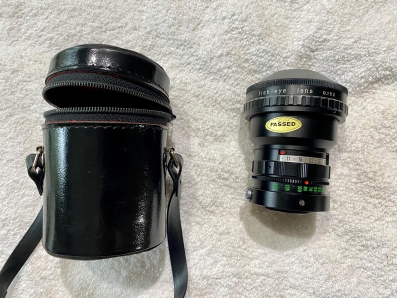 K&ouml;nnte beinhalten: Ein schwarzes Fischaugenobjektiv und eine passende schwarze Tragetasche. Das Objektiv tr&auml;gt den Text "fish-eye lens 0.15x" und einen "PASSED"-Aufkleber. Die Tasche ist zylindrisch mit Rei&szlig;verschluss und Schulterriemen.