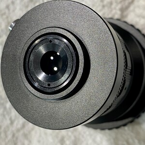 K&ouml;nnte beinhalten: Nahaufnahme eines schwarzen Kameraobjektivs mit strukturierter, matter Oberfl&auml;che. Das Objektiv hat mehrere konzentrische Ringe und eine zentrale Blende mit kleinen, reflektierenden Elementen. Das Gesamtdesign ist elegant und modern.