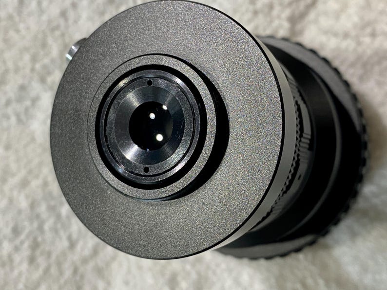 K&ouml;nnte beinhalten: Nahaufnahme eines schwarzen Kameraobjektivs mit strukturierter, matter Oberfl&auml;che. Das Objektiv hat mehrere konzentrische Ringe und eine zentrale Blende mit kleinen, reflektierenden Elementen. Das Gesamtdesign ist elegant und modern.