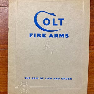May include: A vintage Colt Fire Arms brochure with a tan cover. The blue logo features the word "COLT" above "FIRE ARMS". The text "THE ARM OF LAW AND ORDER" is printed at the bottom. A horse is embossed in the center.