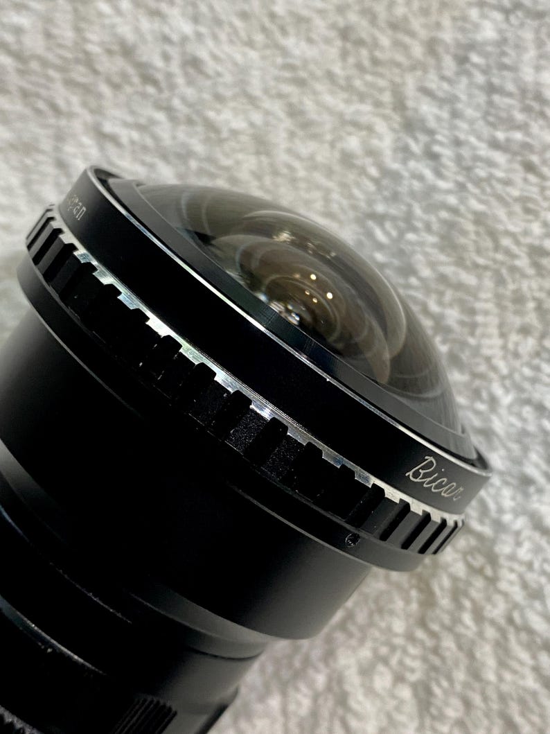 K&ouml;nnte beinhalten: Nahaufnahme eines schwarzen Kameraobjektivs mit einem gew&ouml;lbten Glaselement. Das Objektiv hat einen silbernen Ring mit geriffelter Textur und das Wort "Bicar" ist auf der Seite aufgedruckt. Das Objektiv ist f&uuml;r die Fotografie konzipiert.