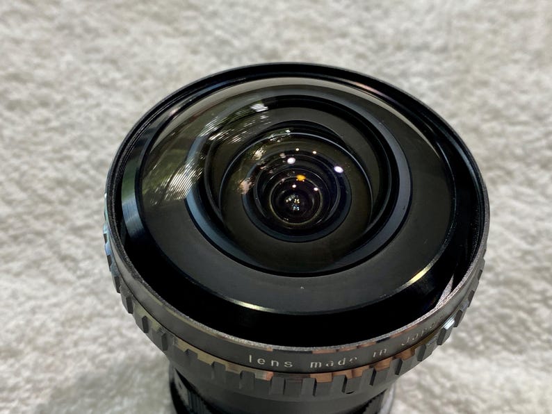 K&ouml;nnte beinhalten: Nahaufnahme eines schwarzen Kameraobjektivs mit Weitwinkeldesign. Das Objektiv hat eine gew&ouml;lbte Glasoberfl&auml;che und einen Metallring mit dem Text "lens made in Japan". Das Objektiv ist f&uuml;r ein breiteres Sichtfeld konzipiert.