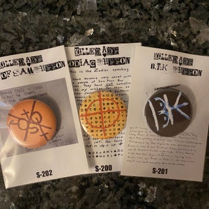 May include: Three button pins with different designs. The first pin is orange and brown with a symbol on it. The second pin is yellow and red with a symbol on it. The third pin is brown with a blue symbol on it. The pins are labeled "Killer Are of Sam Bitton", "Killer Are Zodiac Button", and "Killer Are B.T.K Button".
