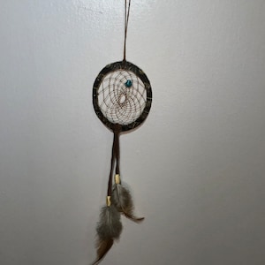 May include: A brown dreamcatcher with a turquoise bead hanging from a brown cord. The dreamcatcher has feathers attached to the bottom.