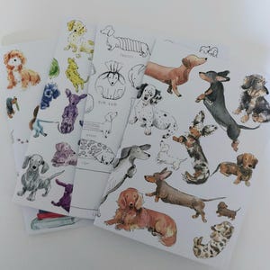 May include: Four different notebooks featuring illustrations of dachshunds in various poses and colours. The notebooks have the words "dim sum", "tiger", "zenna", and "tuxedo" written on them.