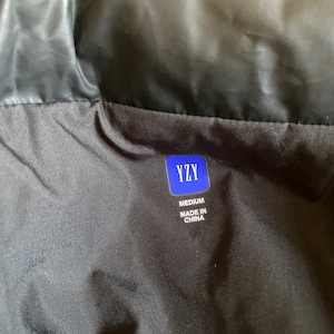May include: Black garment with a label that reads "YZY" in a blue rectangle. Below the logo, the label states "MEDIUM" and "MADE IN CHINA". The garment appears to be made of a shiny, possibly nylon, material.