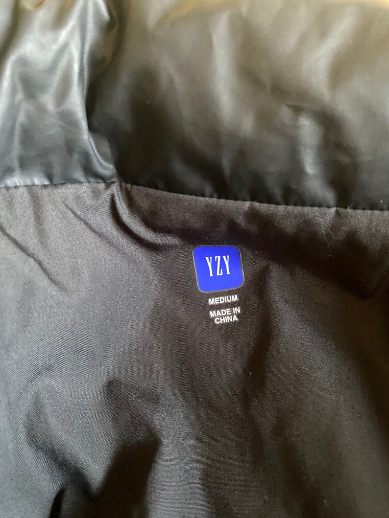 May include: Black garment with a label that reads "YZY" in a blue rectangle. Below the logo, the label states "MEDIUM" and "MADE IN CHINA". The garment appears to be made of a shiny, possibly nylon, material.