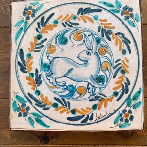May include: Square ceramic tile with a hand-painted design. The tile features a light blue rabbit in a circular blue and gold floral pattern. The border has teal and gold floral accents. The tile is on a wooden surface.