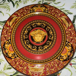 Puede incluir: Un plato decorativo rojo con una cabeza de Medusa dorada en el centro. El plato presenta un borde negro y dorado con adornos de volutas y un borde dorado. Un art&iacute;culo de lujo.
