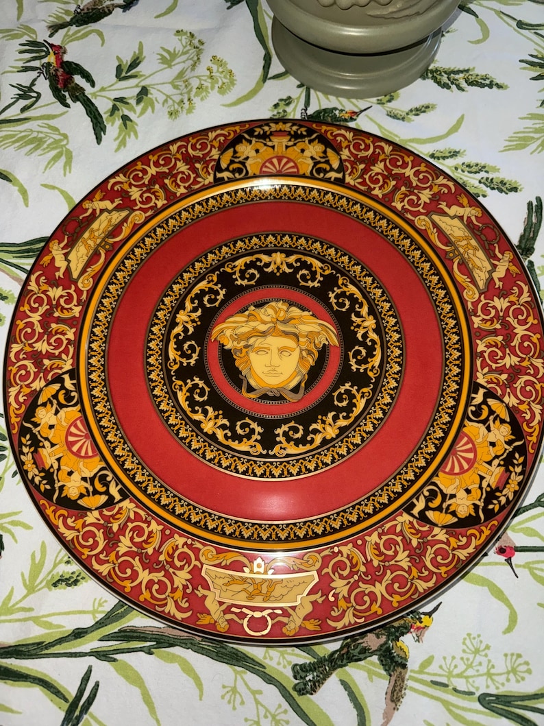 Puede incluir: Un plato decorativo rojo con una cabeza de Medusa dorada en el centro. El plato presenta un borde negro y dorado con adornos de volutas y un borde dorado. Un art&iacute;culo de lujo.