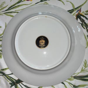 Puede incluir: Un plato de porcelana blanco y gris con el logotipo de Versace en la parte inferior. El plato es redondo con un borde ancho y est&aacute; sobre un fondo con estampado floral.