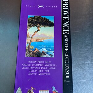 May include: A purple travel guide book with a painting of a Mediterranean scene on the cover. The title is "Provence and the Côte d'Azur" with the text "Knopf Guides" at the top. The book lists cities in Provence and the French Riviera, including Avignon, Nîmes, Arles, Orange, Lourmarin, Marseilles, Aix-en-Provence, Digne, Cannes, Toulon, Biot, Nice, Menton, and Moustiers.