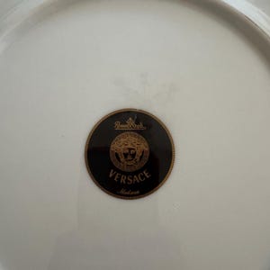 Puede incluir: Plato de porcelana blanca con el logotipo Versace Medusa en negro y dorado. El plato tiene un acabado brillante y un borde ligeramente elevado. Tambi&eacute;n es visible el logotipo de Rosenthal.