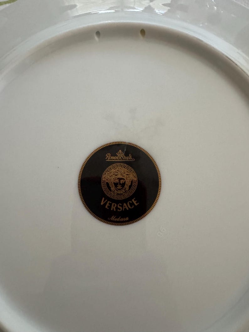 Puede incluir: Plato de porcelana blanca con el logotipo Versace Medusa en negro y dorado. El plato tiene un acabado brillante y un borde ligeramente elevado. Tambi&eacute;n es visible el logotipo de Rosenthal.