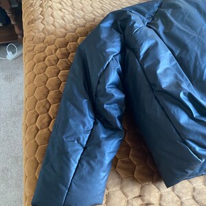 May include: A navy blue puffer jacket, partially visible, with a shiny, smooth texture. The jacket's sleeve is in focus, showing the quilted design and the fabric's sheen. The jacket is a winter outerwear item.