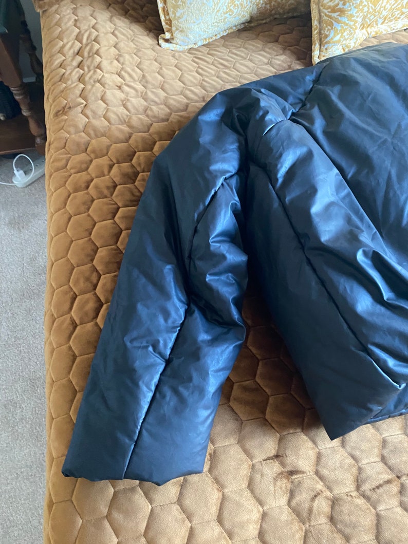 May include: A navy blue puffer jacket, partially visible, with a shiny, smooth texture. The jacket's sleeve is in focus, showing the quilted design and the fabric's sheen. The jacket is a winter outerwear item.