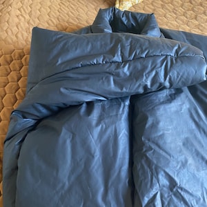May include: A dark gray, puffy jacket with a high collar, lying on a bed. The jacket appears to be made of a smooth, possibly water-resistant material. The bed has a brown quilted cover and two yellow patterned pillows.