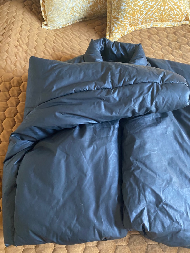 May include: A dark gray, puffy jacket with a high collar, lying on a bed. The jacket appears to be made of a smooth, possibly water-resistant material. The bed has a brown quilted cover and two yellow patterned pillows.