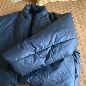 May include: A dark gray puffer jacket with a high collar and long sleeves. The jacket is lying on a brown quilted bedspread, with two decorative yellow pillows in the background.