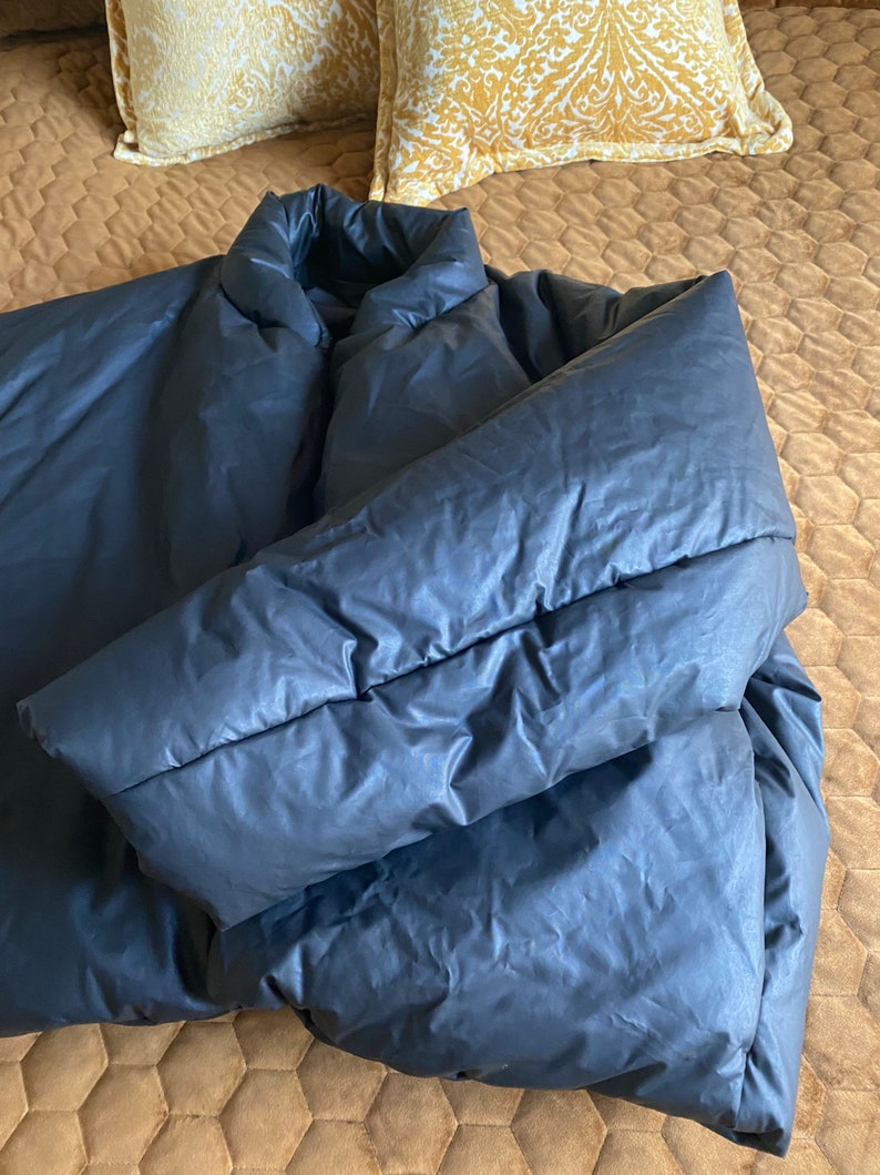 May include: A dark gray puffer jacket with a high collar and long sleeves. The jacket is lying on a brown quilted bedspread, with two decorative yellow pillows in the background.