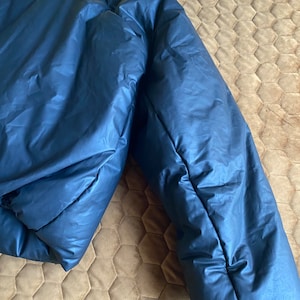 May include: A navy blue puffer jacket, partially visible, with a shiny, smooth texture. The jacket is folded, showing the quilted design and the puffy insulation. The jacket is lying on a brown surface.