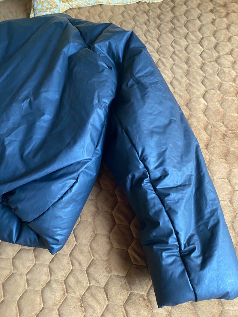 May include: A navy blue puffer jacket, partially visible, with a shiny, smooth texture. The jacket is folded, showing the quilted design and the puffy insulation. The jacket is lying on a brown surface.