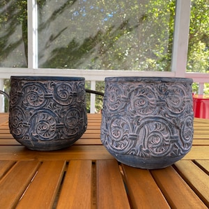 May include: Two dark charcoal gray planters with a rounded shape and a textured, embossed pattern of repeating circular scrolls and foliage. The surface of the planters features a weathered, matte finish with lighter gray highlights in the crevices.